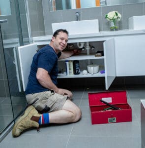 Local plumber for Macleod kneeling down, diligently working to clear a blockage under a sink in Victoria.