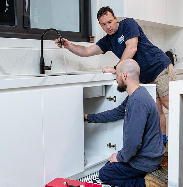 A friendly plumber is fixing a leaking tap, while another kneels down to inspect a blocked drain in a cabinet inside a Victoria home.