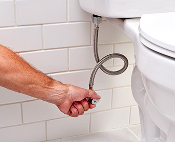 A plumber's hand attaching a flexible water supply line to the bottom of a toilet tank, with clean white subway tiles in the background.