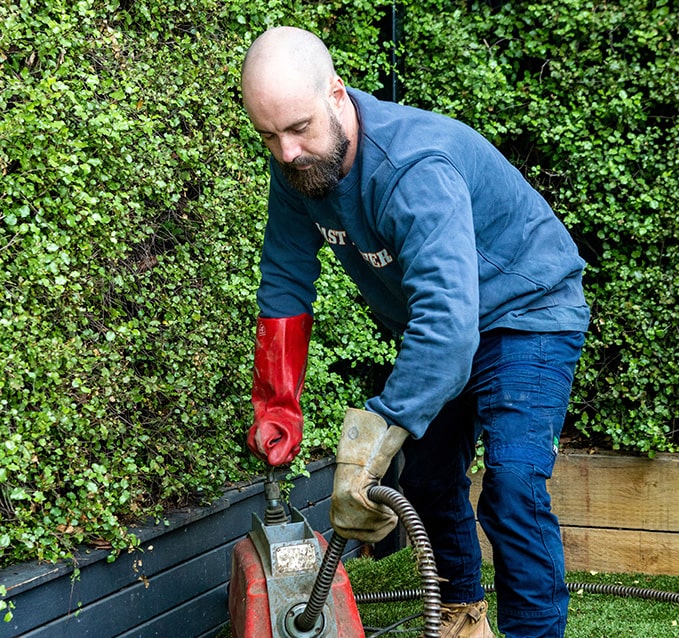 A focused plumber in a blue sweatshirt and red gloves operates a drain snake machine outdoors, with lush greenery in the background.