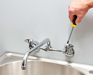 A hand using a yellow and black screwdriver to adjust the hardware on a polished chrome kitchen faucet, set above a gleaming stainless-steel sink against a simple grey wall.
