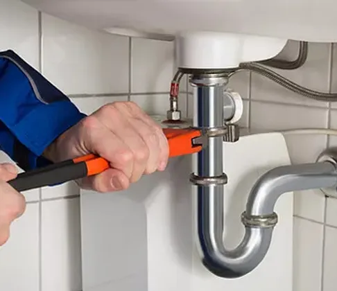 Close-up of a plumber's hands using orange pliers to tighten the connection on a chrome P-trap under a sink, against a backdrop of white tiles.