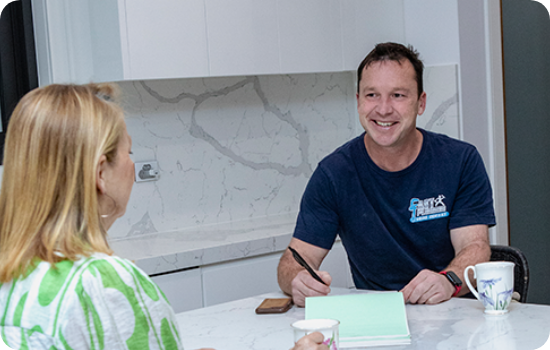 One of the owners of a plumbing business, identifiable by his branded shirt, discussing plumbing needs with a client across a table, with a modern kitchen backdrop featuring marble countertops.